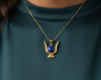 May include: A gold-colored pendant necklace featuring a blue gemstone. The pendant has a unique design with curved elements and a circular blue stone at the center. The necklace is worn against a teal-colored top.