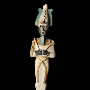 Puede incluir: Estatua detallada de una figura egipcia, con rostro negro y elaborada tocado. La figura viste una prenda blanca con detalles dorados y tiene los brazos cruzados. La estatua está montada sobre una base rectangular.
