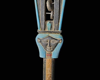 May include: A blue and gold wooden sistrum with a carved female face and hieroglyphs. The sistrum has a rectangular frame with a series of small metal rings inside.