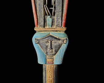 May include: An Egyptian sistrum, a percussion instrument, featuring a carved face with blue hair and a detailed hieroglyphic inscription. The instrument is primarily black with red, blue, and gold accents.