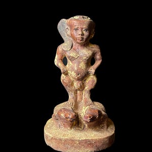 May include: A terracotta figurine of a standing figure with a headdress, holding a staff and standing on two crouching figures. The figurine is painted in shades of brown and yellow.