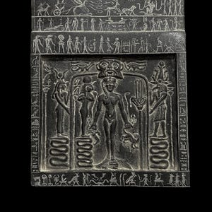 May include: Black and white stone relief carving of an Egyptian goddess standing between two other figures. The goddess is nude and has a headdress with a sun disk and uraeus. The figures on either side of her are holding staffs and wearing traditional Egyptian clothing.