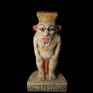 Puede incluir: Estatua detallada, blanquecina, de una deidad egipcia con una cabeza grande, boca abierta y lengua roja. La estatua tiene una cofia amarilla, orejas rojas y un taparrabos rojo y dorado. La base está inscrita con jeroglíficos. Fondo negro.