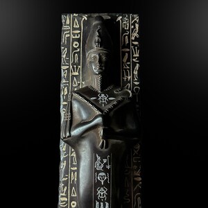 Puede incluir: Una escultura vertical oscura de un antiguo faraón egipcio. La figura se representa con una alta tocado, rasgos faciales detallados y brazos cruzados. Jeroglíficos están tallados a los lados de la escultura. La estatua es de color oscuro, posiblemente negro.
