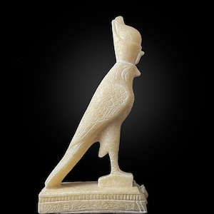 Puede incluir: Una estatua de alabastro blanco del dios egipcio Horus, representado como un halcón con cabeza humana que lleva una corona. La estatua está de pie sobre una base con jeroglíficos.