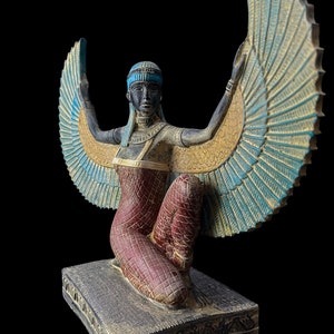 Puede incluir: Una estatua alada de una diosa egipcia con los brazos extendidos, arrodillada sobre una base. La estatua est&aacute; pintada en tonos dorados, azules, rojos y negros.