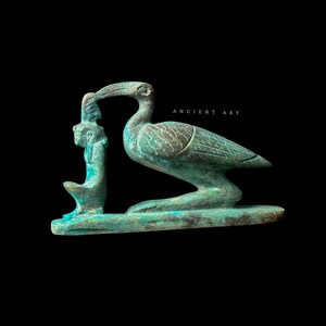 Puede incluir: Una escultura egipcia antigua de color turquesa de un pájaro sosteniendo una figura. La escultura está sobre un fondo negro. El texto "ANCIENT ART" está en blanco en la parte superior derecha.
