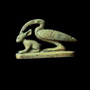 Puede incluir: Una escultura tallada de un conejo y un pájaro, posiblemente un ibis, en color marrón claro. El conejo está sentado y el pájaro está a su lado. La escultura está sobre una base rectangular sobre un fondo negro.
