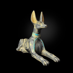 Pode incluir: Uma estátua de Anúbis preta e dourada com detalhes azuis e dourados. A estátua é de um chacal com a cabeça virada para o lado. A estátua está deitada com as patas esticadas.