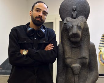 May include: A man stands next to a dark gray statue of a lion-headed figure. The statue has a large sun disc above its head. The man wears a black jacket, a watch, and headphones around his neck. The background is a neutral-toned wall.