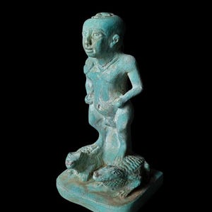 May include: A turquoise-colored statue of a standing figure with a shaved head, holding its sides. The figure stands on a base with two crocodile figures. The statue has a weathered appearance and is set against a black background.