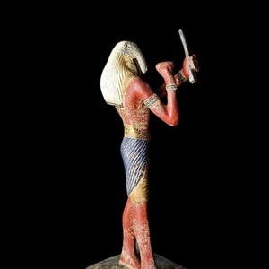 Puede incluir: Estatua detallada de una deidad egipcia, con cabeza de pájaro y largo pico, con tocado blanco. La figura tiene piel roja, detalles azules y dorados y sostiene una cuchilla. La estatua está sobre un fondo negro.