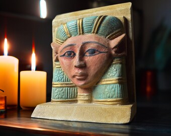 Puede incluir: Un sujetalibros decorativo con la cara de un faraón egipcio. El sujetalibros está pintado en tonos beige, turquesa y dorado. La cara del faraón tiene ojos oscuros y detalles rojos. El sujetalibros es rectangular y se mantiene en posición vertical.