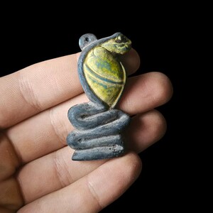 Op de afbeelding: Een kleine, gestileerde cobra-hanger met een lus om op te hangen. De kop van de cobra is goudkleurig en het lichaam is donkerblauw-grijs. De hanger wordt in een hand gehouden tegen een zwarte achtergrond.