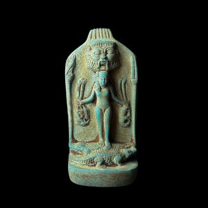May include: A turquoise blue Egyptian amulet depicting a goddess standing on two crocodiles. The goddess is holding two staffs with a snake head on top. The amulet has a carved face of a god at the top.