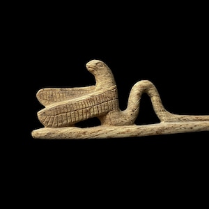 Op de afbeelding: Een houten beeldje van een vogel met uitgespreide vleugels, zijn kop gedraaid naar de zijkant. De vogel zit op een lange, dunne houten stok.