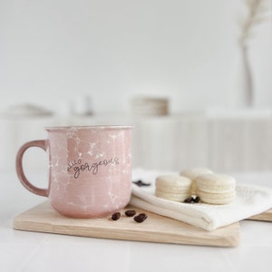 May include: Pink ceramic mug with a white marbled design and the text "Hello Gorgeous" in black. The mug is sitting on a wooden cutting board with coffee beans scattered around it.