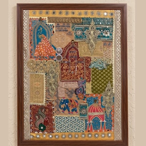Op de afbeelding: Een rechthoekig wandkunstwerk in een donkerbruine lijst met een collage van geborduurde stoffen lappen. Het textielontwerp bevat een pauw, een olifant en architecturale motieven in levendige tinten goud, rood, blauw en groen.
