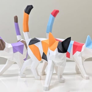 Könnte beinhalten: Drei weiße Katzenfiguren mit geometrischen Designs. Jede Katze hat ein anderes Farbschema, darunter Orange, Schwarz und Weiß; Lila, Braun und Weiß; und Rot, Blau und Weiß. Die Katzen stehen auf einer weißen Oberfläche.
