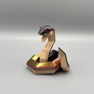 Könnte beinhalten: Eine geometrische Papierskulptur einer Schlange. Die Schlange hat einen beigefarbenen Körper, einen schwarzen und roten Kopf und goldene, rote und schwarze Akzente. Die Schlange ist aufgerollt und aufrecht, mit erhobenem Kopf.