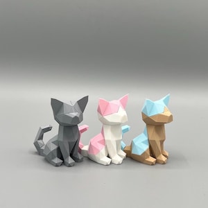 Könnte beinhalten: Drei geometrische Katzenfiguren, eine graue, eine weiße mit rosa Akzenten und eine braune mit blauen Akzenten.