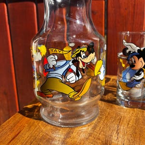 Pu&ograve; includere: Una caraffa di vetro trasparente e un bicchiere con personaggi Disney. La caraffa ha un'immagine a cartoni animati di Pippo che tiene un cartello con scritto "YAY!". Il bicchiere ha un'immagine di Topolino. Entrambi sono su una superficie di legno.