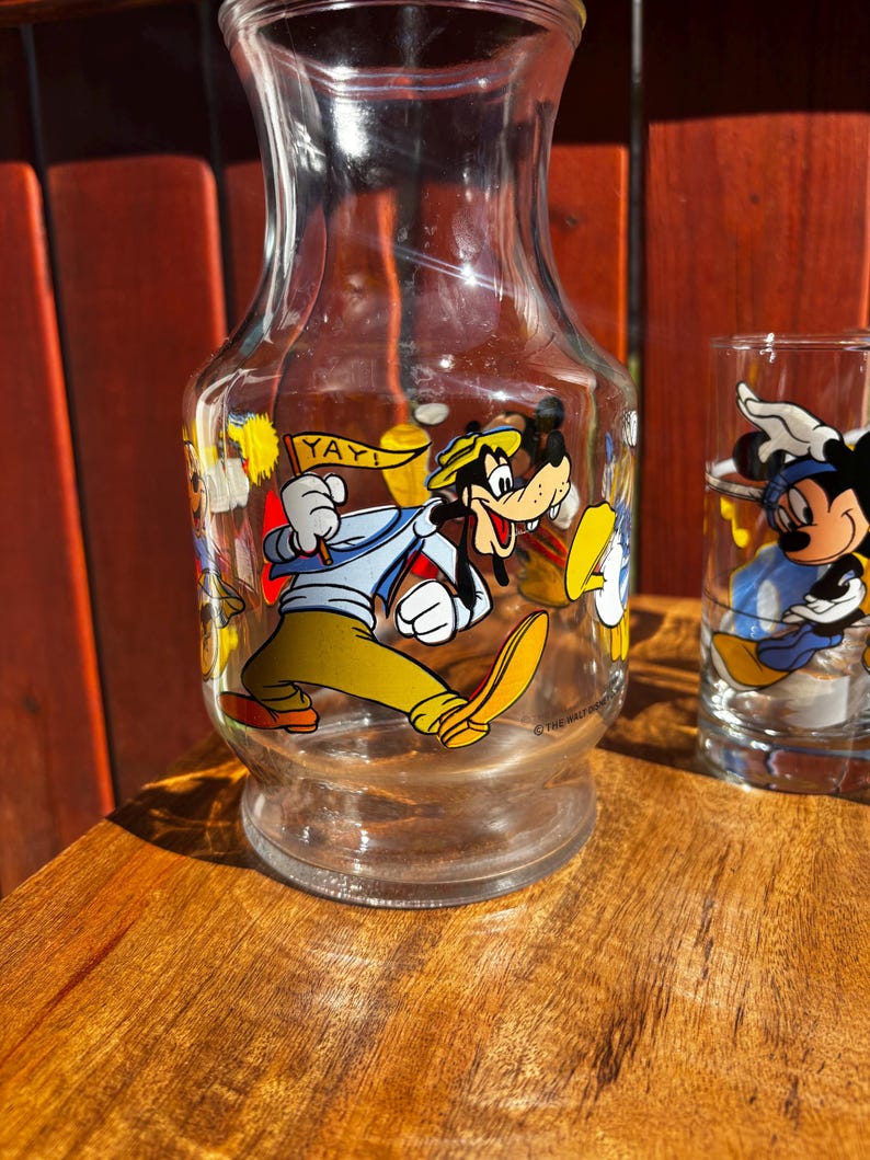 Pu&ograve; includere: Una caraffa di vetro trasparente e un bicchiere con personaggi Disney. La caraffa ha un'immagine a cartoni animati di Pippo che tiene un cartello con scritto "YAY!". Il bicchiere ha un'immagine di Topolino. Entrambi sono su una superficie di legno.