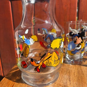 Pu&ograve; includere: Una caraffa di vetro trasparente e un bicchiere con disegni di personaggi dei cartoni animati. La caraffa presenta Pluto, Paperino e altri personaggi. Il bicchiere ha Topolino. Il set &egrave; su una superficie di legno.