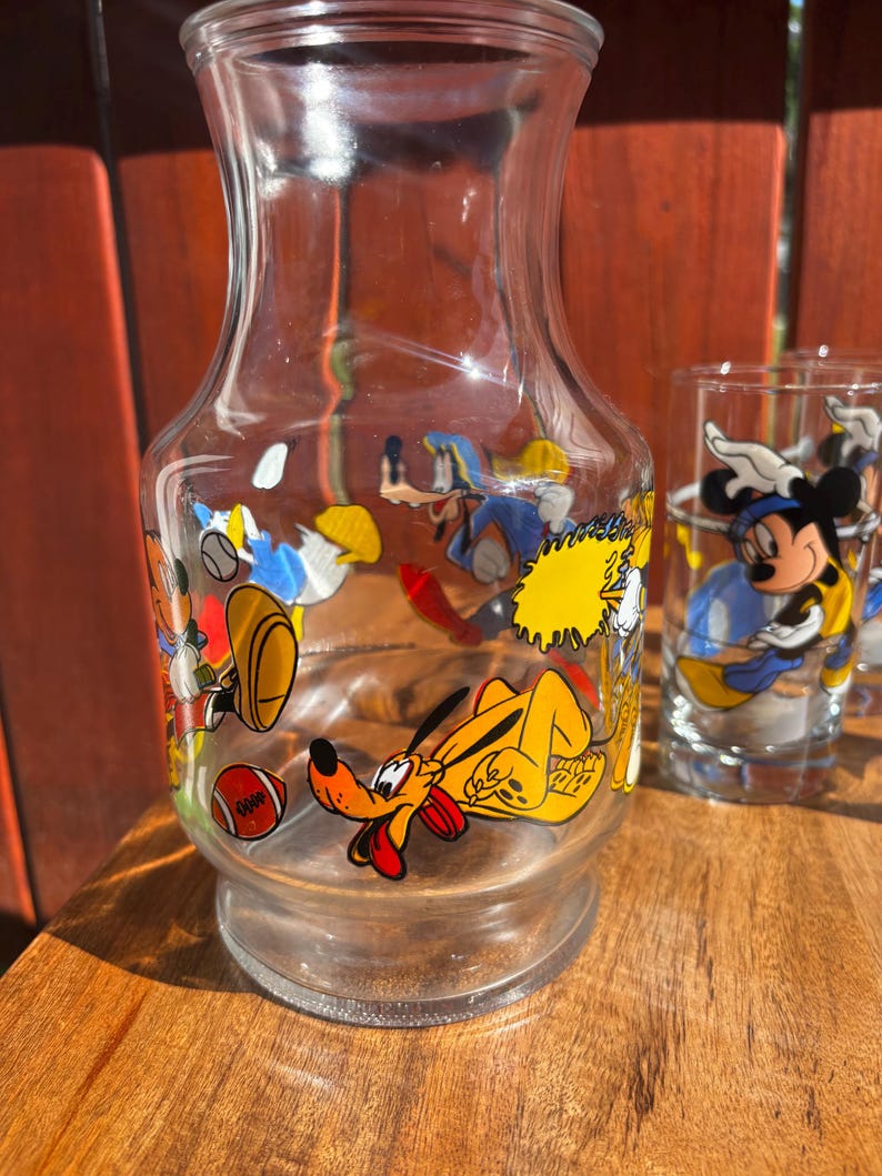 Pu&ograve; includere: Una caraffa di vetro trasparente e un bicchiere con disegni di personaggi dei cartoni animati. La caraffa presenta Pluto, Paperino e altri personaggi. Il bicchiere ha Topolino. Il set &egrave; su una superficie di legno.