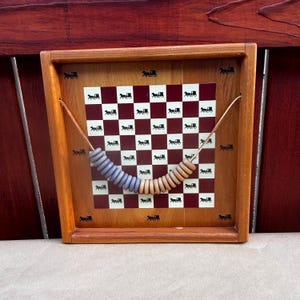 May include: A wooden abacus with a burgundy and white checkered background. The background is decorated with a repeating pattern of a horse-drawn carriage. The abacus has two rows of wooden beads in shades of blue and tan.