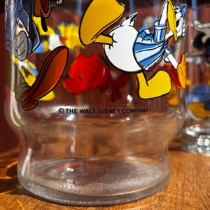 Pu&ograve; includere: Bicchiere trasparente con grafica di personaggi dei cartoni animati, tra cui Paperino e Pippo. La base del bicchiere &egrave; visibile e il testo "THE WALT DISNEY COMPANY" &egrave; stampato sul bicchiere.