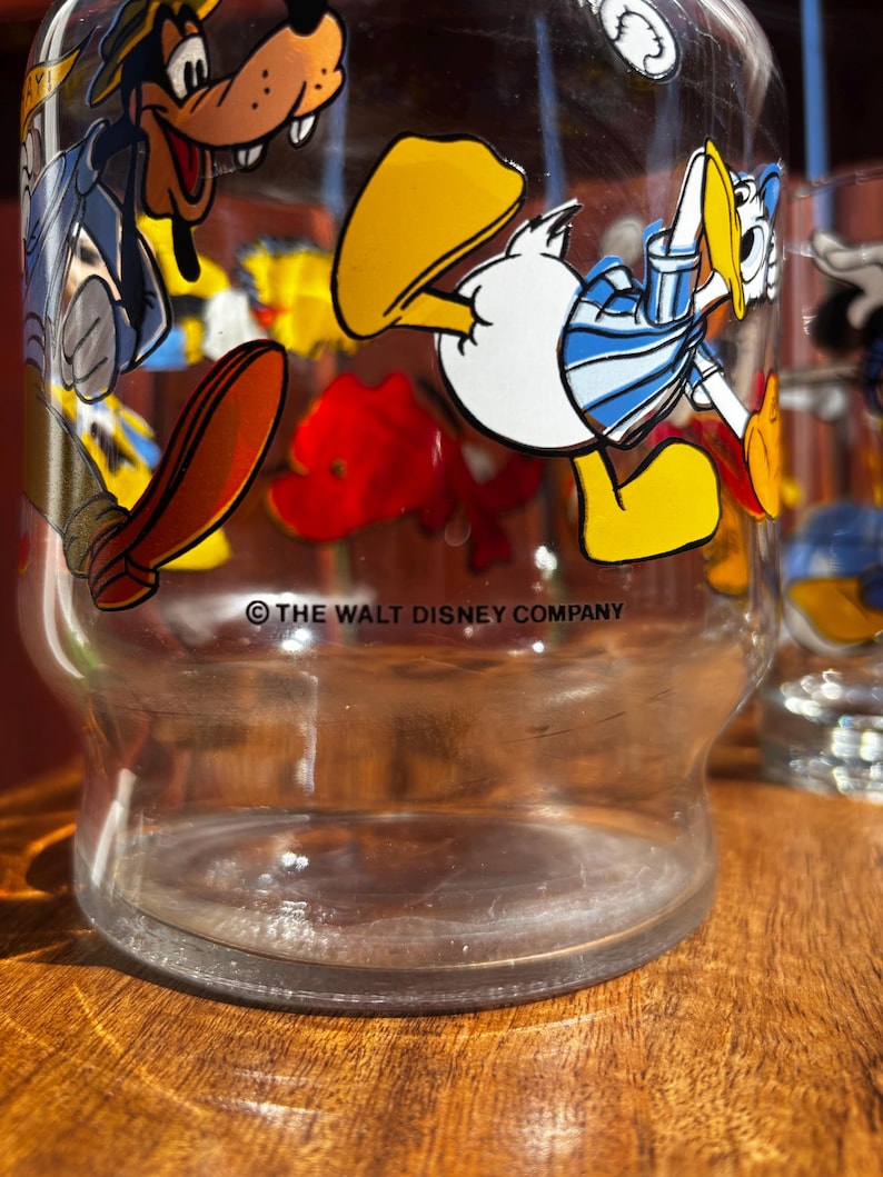 Pu&ograve; includere: Bicchiere trasparente con grafica di personaggi dei cartoni animati, tra cui Paperino e Pippo. La base del bicchiere &egrave; visibile e il testo "THE WALT DISNEY COMPANY" &egrave; stampato sul bicchiere.