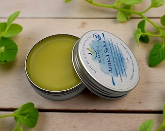 May include: A round silver metal tin containing a smooth olive-green arnica salve. The lid is placed beside the open tin, featuring a white label with blue text that reads Botanical Arnica Salve, 2 oz., and a list of natural ingredients.