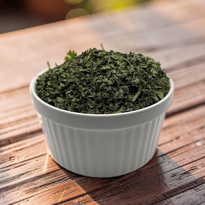 May include: A white ceramic ramekin with vertical fluted sides filled to the brim with a mound of dried, finely chopped dark green herbs. The bowl has a smooth, matte finish and a wide, circular opening, containing a heap of textured plant matter.