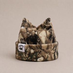 May include: A brown, black, and tan camouflage patterned fleece beanie with two pointed ears at the top. The hat features a folded brim and a small white rectangular tag on the front with the black text KEANIES and a graphic of a crocodile.