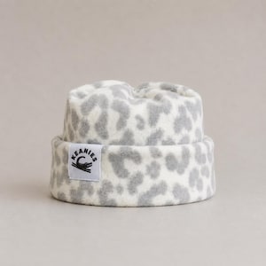 May include: A white and gray leopard print beanie with a folded cuff. A white rectangular fabric label is sewn onto the front of the cuff, featuring the black text KEANIES and a graphic illustration of a crocodile tail.