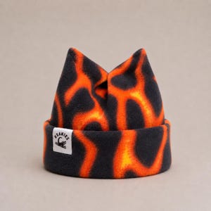 May include: A cuffed beanie featuring a vibrant orange and black lava-like pattern. The hat has two pointed top corners and a small white rectangular label on the front cuff with the text KEANIES and a graphic of a small crocodile.