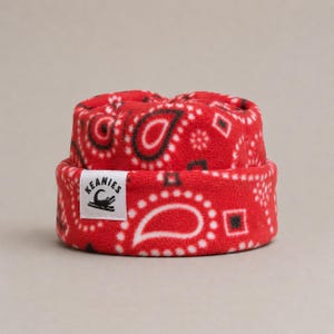 May include: A red fleece beanie with a white, black, and red paisley pattern. The hat features a folded cuff with a white rectangular label sewn on the front. The label displays the text KEANIES above a black graphic of a crocodile silhouette.