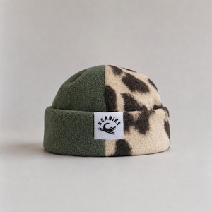 May include: A fleece beanie featuring a split design with one half in solid olive green and the other half in a beige and dark brown leopard print pattern. A white rectangular patch on the front displays the text KEANIES with a crocodile graphic.