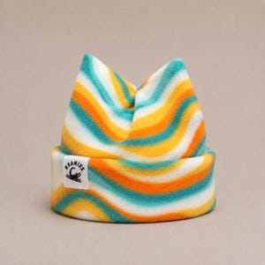 May include: A fleece beanie featuring a wavy, multi-colored pattern of teal, orange, and white stripes. The hat has a folded cuff with a white rectangular label sewn on the front that displays the text KEANIES and a small crocodile graphic.