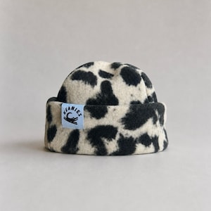 May include: A cuffed beanie featuring a black and cream cow-print pattern. A light blue rectangular tag is sewn onto the front cuff, displaying the text KEANIES and a graphic illustration of a crocodile silhouette in black ink.