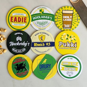 以下が含まれることがあります： 様々なデザインとテキストが施された9つのビールコースターのコレクション。コースターには、「Eadie」、「Hoolahan's」、「Holt Lager」、「Huckerby's」、「Munich 93」、「Pukki」、「Iwan Roberts」、「Drury」、および「Gunn & Son Brewing」などのブランドが表示されています。コースターは丸く、主に黄色、緑、白です。