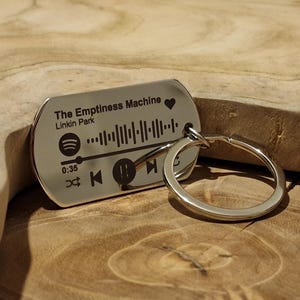 Könnte beinhalten: Ein silberfarbener Metall-Schlüsselanhänger mit einem Audio-Wellenform-Design im Spotify-Stil. Der Anhänger trägt den eingravierten Text "The Emptiness Machine" von Linkin Park, Wiedergabesteuerung und eine Zeitanzeige.