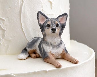 May include: A small, detailed figurine of a merle-colored dog with grey, white, and tan fur, lying down in a resting position. The dog figurine is placed on the side of a two-tiered white cake with a textured, swirled frosting finish.
