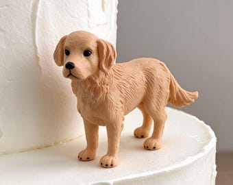 May include: A small, light brown golden retriever figurine standing on a white, multi-tiered cake. The dog figure has textured fur, a black nose, and dark eyes, positioned on the smooth, white frosted surface of the cake tier.