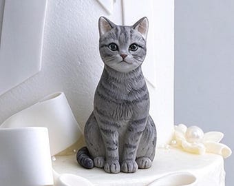 May include: A gray and white tabby cat figurine with dark stripes, pointed ears, and large eyes, positioned on top of a white cake decorated with large white fondant bows and small white pearl accents.