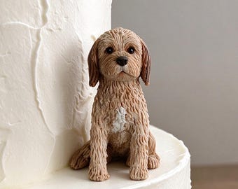 May include: A small, light brown, curly-haired dog figurine sits atop a two-tiered white cake. The dog has a white patch on its chest, dark eyes, and a black nose. The cake features a textured, swirled white frosting finish throughout.