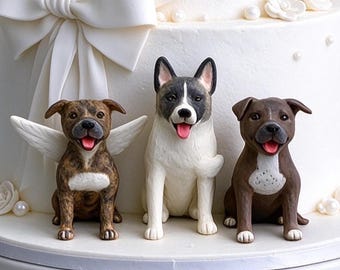 May include: Three dog figurines displayed on a white cake. The left dog is brindle with white wings, the center dog is black and white, and the right dog is brown with a white chest patch. All three dogs have open mouths with pink tongues.
