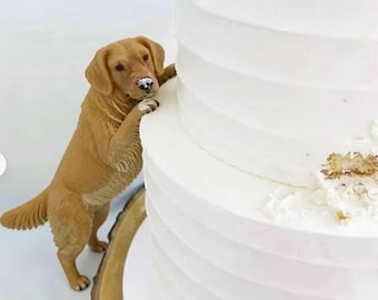May include: A golden retriever figurine standing on its hind legs with its front paws resting against the side of a multi-tiered white cake. The dog has light brown fur and a dark nose, while the cake features smooth white frosting.