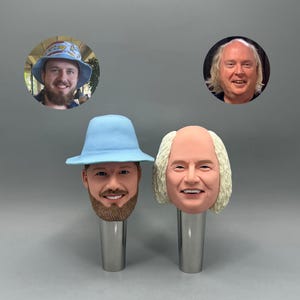 May include: Two novelty bottle stoppers featuring sculpted faces. One has a light blue hat and a beard, the other has white hair. Both are mounted on silver-colored metal stems. The background includes two circular portraits.